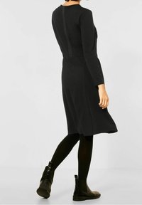 Svart långärmad klänning med figursydd överdel, utsvängd kjol och texturerad tyg. Har en vertikal dragkedjedetalj på baksidan.