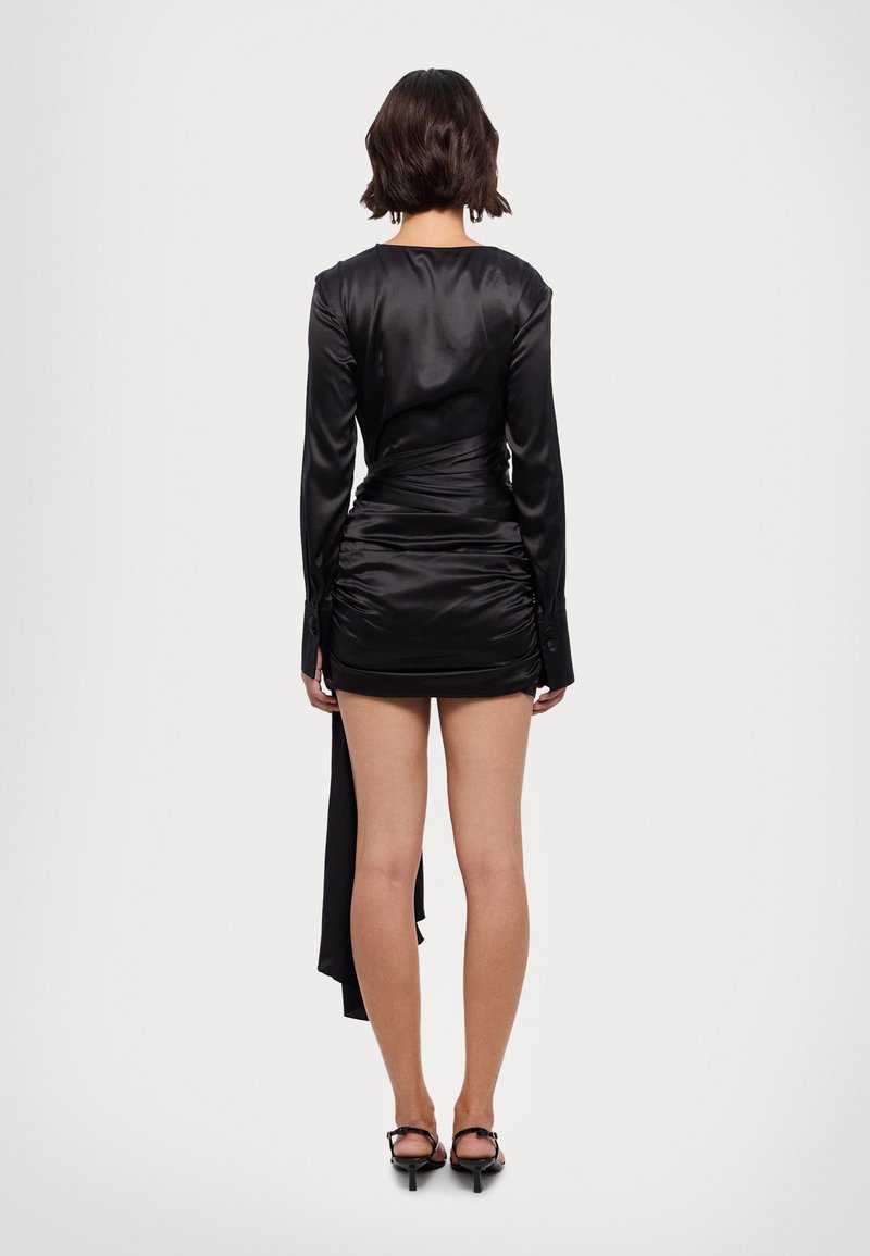 Vestido de satén negro con mangas largas, fruncido en la parte posterior, dobladillo asimétrico y puños abotonados, combinado con sandalias de tacón negras.
