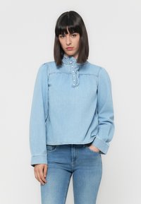 Γυναίκα με ίσια σκούρα μαλλιά, φορώντας ένα ανοιχτό μπλε denim πουκάμισο με ρέλι και μακριά μανίκια, καθώς και μπλε τζιν, όρθια μπροστά σε λευκό φόντο.