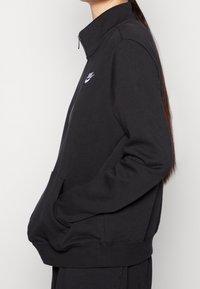 Svart zip-up sweatshirt med ståkrage, tillverkad av mjukt material. Har en liten vit Nike-logotyp på vänster bröst och framfickor.