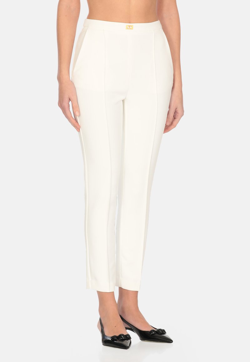Pantaloni bianchi su misura con una texture liscia, caratterizzati da una vestibilità slim e un dettaglio in metallo dorato in vita. Abbinati a scarpe nere a punta.