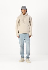 Felpa in pile beige con tasca frontale e coulisse, abbinata a jeans larghi blu chiaro e sneakers crema. Cappellino grigio chiaro.