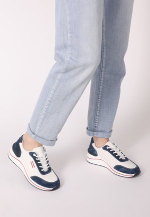 White and navy sneakers with blue suede accents, lace-up design, rounded toe, and red stripe on the sole, paired with rolled-up light blue jeans.