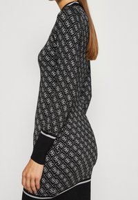 Pull en tricot noir avec un motif géométrique blanc, col rond et poignets noirs contrastants. La texture semble lisse.