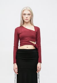 Burgundski top s dugim rukavima, s omotanim dizajnom i bočnim čvorom, uparen s crnom uskom suknjom s detaljima na vezanje i teksturiranom tkaninom.