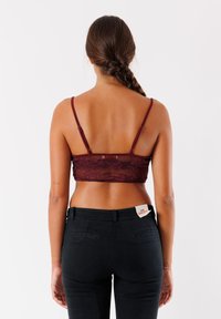 Bralette in pizzo borgogna con spalline regolabili, costruzione semi-trasparente, abbinato a pantaloni neri a vita alta con etichetta sul cinturino.
