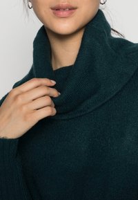 Maglioncino a collo alto in tonalità verde acqua, realizzato in materiale lavorato a maglia, caratterizzato da un colletto arrotolato e da una mano che sfiora delicatamente il tessuto.