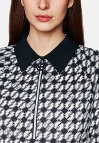 Schwarzes und weißes Zip-up-Oberteil mit Kragen, geometrischem Muster und Branding-Details, aus einem glatten Stoff gefertigt.