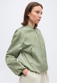 Žena u svijetlozelenoj bomber jakni na patentni zatvarač s rukama u džepovima, gleda udesno, na jednostavnoj svijetloj pozadini.