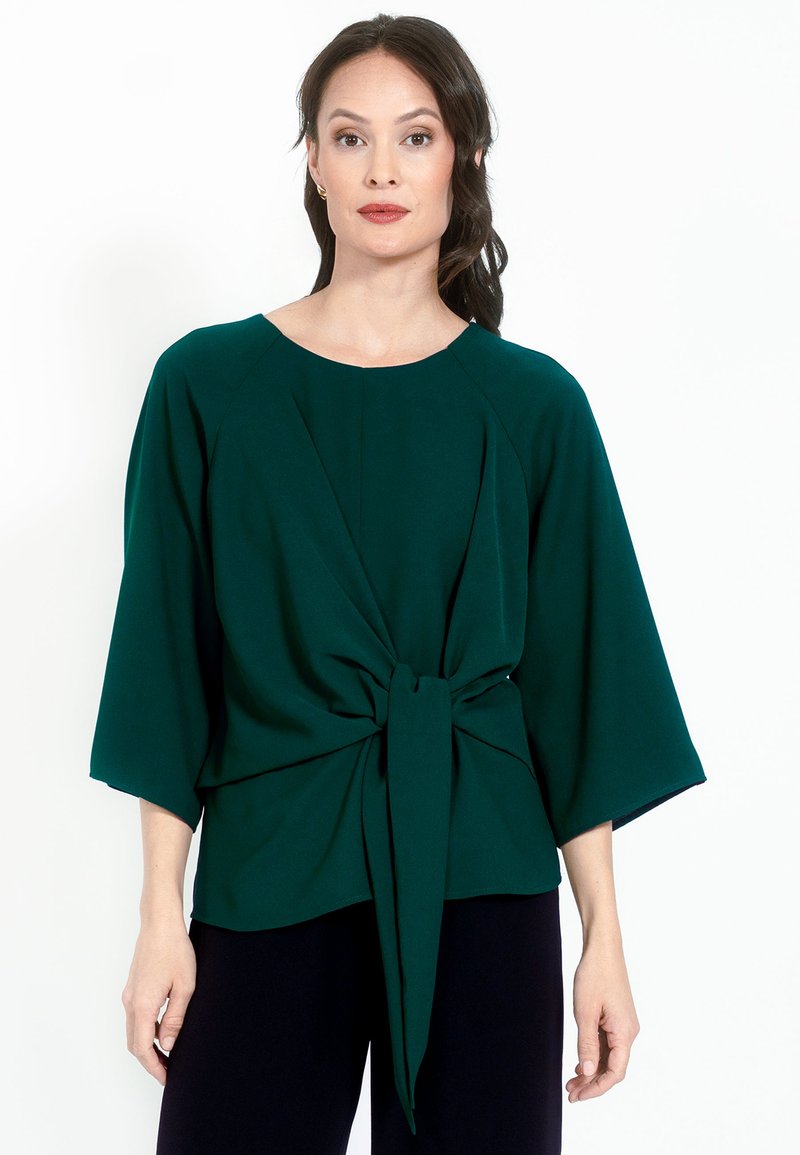 Blusa verde con maniche ampie e dettaglio a nodo frontale. Realizzata in tessuto morbido; presenta un collo rotondo e pieghe sottili.
