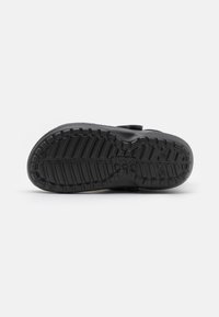 Semelle de chaussure noire en caoutchouc texturé, avec des rainures profondes pour l'adhérence, et un design épuré, comportant un détail de logo circulaire près du talon.