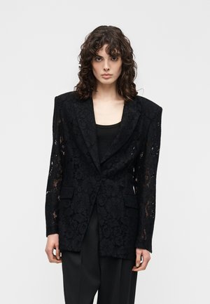 Woman with curly dark hair wearing a black lace blazer with padded shoulders and black pants, standing against a plain light background.