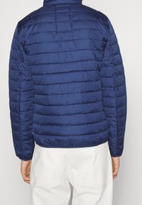 Donkerblauwe doorgestikte pufferjas met horizontale naden, een hoge kraag en elastische manchetten. De stof lijkt glad en lichtgewicht.