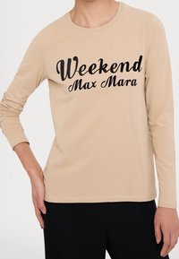 Beige Langarmshirt mit schwarzem "Weekend Max Mara"-Schriftzug, getragen von einer Person mit der Hand auf der Hüfte, kombiniert mit schwarzen Hosen.