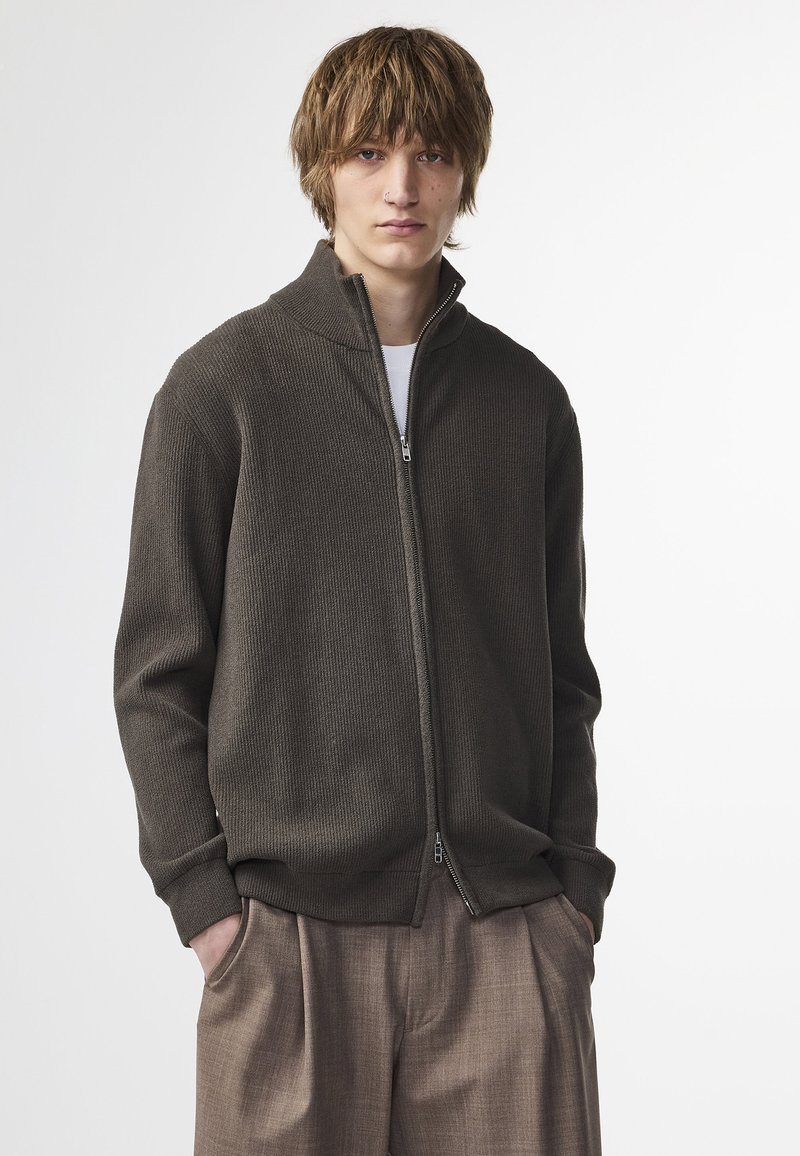Veste zippée en tricot marron avec un col montant et une texture côtelée, associée à un pantalon plissé beige. Poches avant visibles.