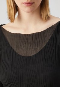 Svart plisserad klänning med en genomskinlig, texturerad krage. Tyget har vertikala linjer och en scalloped kant längs kragen.