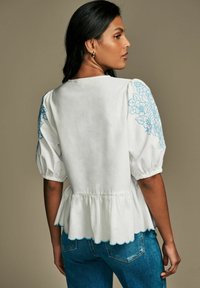 Camicia bianca con maniche a sbuffo, ricami floreali blu sulle spalle, orlo a peplum, bordi ondulati, indossata con jeans blu.