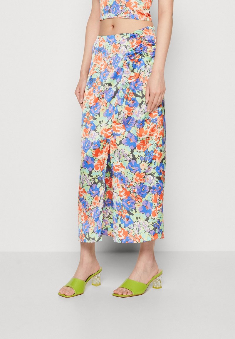 Jupe portefeuille fleurie présentant un mélange de fleurs orange, bleues et vertes sur un fond sombre, avec une fente haute sur la cuisse et une texture douce. Sandales à talons vertes.