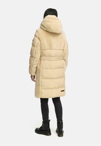 Beige gewatteerde jas met een pluizig, vachtachtig bovenstuk en een afneembare capuchon. Heeft een getailleerde taille en een soepele textielafwerking.