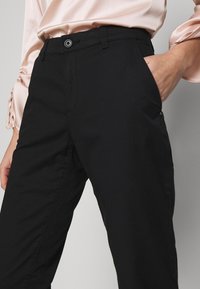Pantalon noir à texture lisse, doté d'une fermeture par bouton, de poches avant et d'une coupe ajustée, associé à un chemisier rose clair.