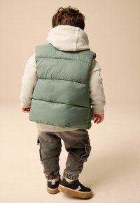 Grüne gefütterte Weste über einem hellgrauen Hoodie, kombiniert mit dunkelgrauen Cargo-Hosen und schwarzen Sneakers. Weicher Stoff, lockere Passform, lässiges Design.