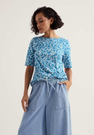 Mujer con cabello corto y rizado, vistiendo una blusa de manga corta azul con estampado y pantalones sueltos de mezclilla clara, con la mano en el bolsillo.