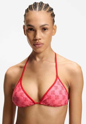 Mujer con el cabello trenzado que lleva un top de bikini triangular rosa y rojo con un patrón repetido de texto "hugo".