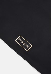 Černá látková taška s obdélníkovou zlatou logovou destičkou s vyraženým nápisem "even&odd" na hladkém povrchu. Jednoduchý, minimalistický design.