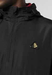 Veste noire légère avec un col montant, une fermeture éclair à l'avant, et un logo triangulaire en noir et or. Texture lisse, design minimaliste.