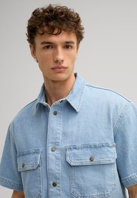 Camicia di jeans azzurro chiaro a maniche corte con colletto a punta, due tasche sul petto con chiusura a bottone e una finitura leggermente texturizzata.