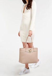 Sac fourre-tout beige avec un accent en logo doré, forme structurée et deux poignées. Conçu avec une texture lisse et des détails matériels minimalistes.
