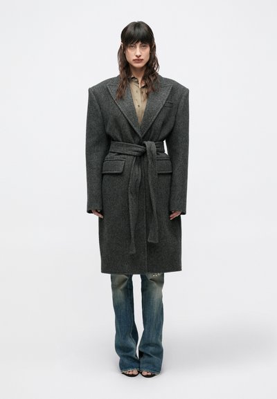 Long manteau en laine gris avec de larges revers, ceinture et poches latérales, porté sur une chemise claire à boutons et un jean bleu usé.