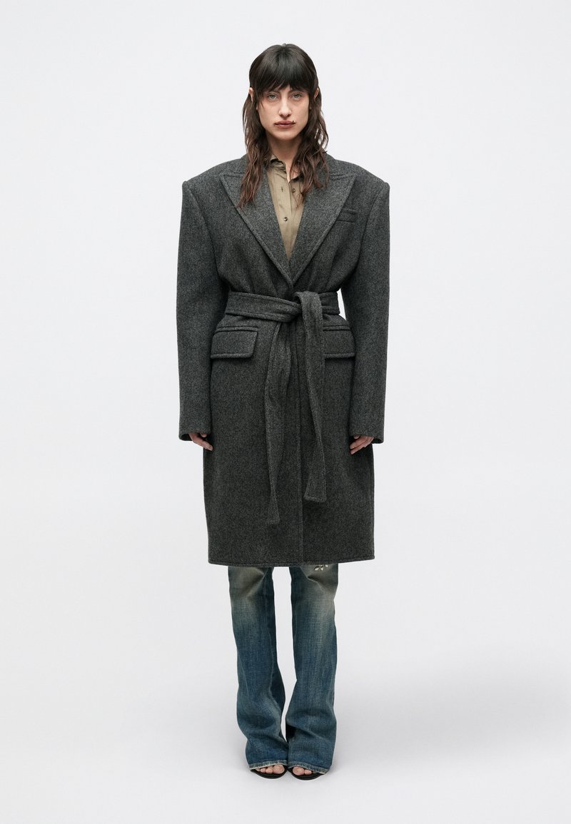 Lang grå uldfrakke med brede revers, bælte og sidelommer, båret over en lys skjorte med knapper og slidte blå jeans.