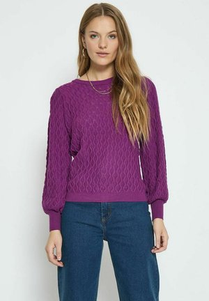 Pull en tricot violet avec un motif en losanges, col rond et manches longues légèrement bouffantes. Associé à un jean bleu.