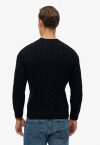 Marineblauwe kabelgebreide trui met lange mouwen, voorzien van een ronde halslijn en ribgebreide boorden. Draagt de trui met lichtblauwe jeans.