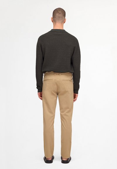 Homme de dos portant une chemise à manches longues sombre avec texture, un pantalon beige et des chaussures noires sur un fond blanc uni.