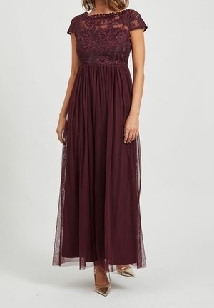 Femme portant une robe bordeaux longue jusqu'au sol, à manches courtes, corsage en dentelle avec superposition transparente, associée à des escarpins dorés à bout pointu.