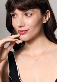 Złoty naszyjnik o zakrzywionym kształcie z małymi, przezroczystymi kamieniami szlachetnymi. Noszony z bluzką w czarno-białe paski. Gładka tekstura skóry, naturalny makijaż.