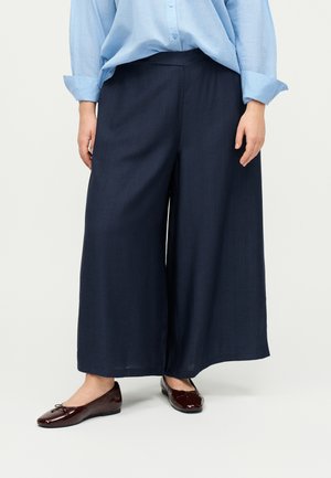 Personne portant un pantalon large bleu marine ample, une chemise bleu clair boutonnée avec manches retroussées et des ballerines marron foncé.