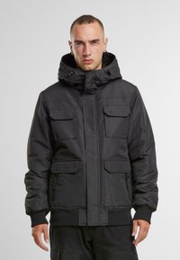 Geacă neagră cu glugă, prevăzută cu un fermoar frontal, multiple buzunare, manșete ribbate și un material texturat. Designul oferă un aspect structurat și practic.