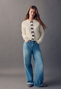 Weißer Strickcardigan mit schwarzen Schleifenknöpfen, kombiniert mit hoch taillierten, weit geschnittenen blauen Jeans und verzierten schwarzen spitzen Schuhen.