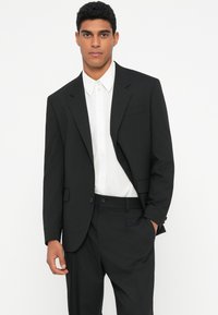 Traje negro con un diseño a medida, que presenta una chaqueta de un solo botón y pantalones de frente plano, combinados con una camisa blanca de botonadura.