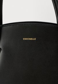 Zwarte leren handtas met gouden "COCCINELLE"-logo, gecentreerd nabij de bovenkant, en twee zwarte handvatten zichtbaar.