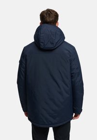 Veste isolante bleu marine avec capuche, présentant une texture lisse, une forme allongée et des poignets élastiques. Vue de dos montrant un design simple.