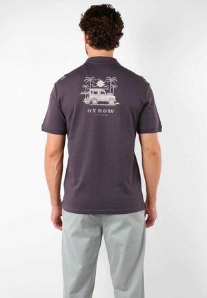 T-shirt gris foncé avec un graphique décoloré d'un van et de palmiers dans le dos. Manches courtes et tissu doux, associé à un pantalon gris clair.
