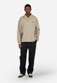 Bež majica s valovitom pletenico in dolgimi rokavi, s ovratnikom in tremi gumbi. V kombinaciji z črnimi hlačami, ki imajo majhen logotip. Črne superge z belimi podplati.