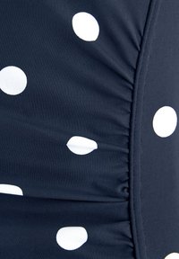Mørkeblå svømmedrakt med hvite prikker; har rynker langs siden; laget av elastisk, glatt stoff som gir en tettsittende fasong.