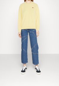 Sudadera amarilla clara con cuello redondo y pequeño logotipo rojo, combinada con unos vaqueros de pierna ancha azules con costuras diagonales y zapatillas blancas.