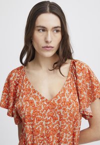 Blommig v-ringad topp med korta, sväva ärmar. Basfärg beige med ett abstrakt blommönster i orange och rött. Lättviktsmaterial, avslappnad passform.