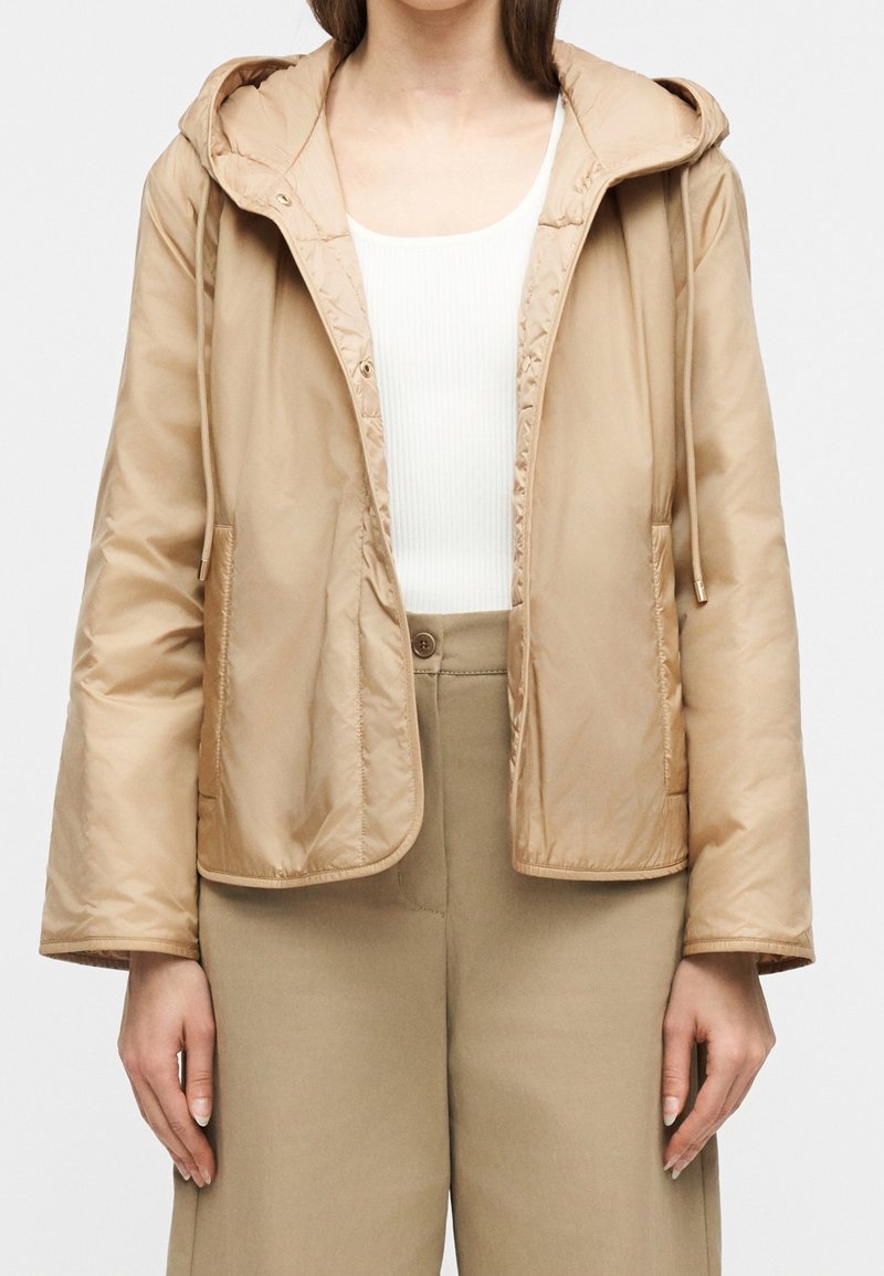 Femme portant une veste à capuche beige sur un haut côtelé blanc et un pantalon beige taille haute sur fond uni.